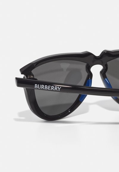 Burberry saulės akiniai su juodu plastiku rėmu, pilkais objektyvais ir mėlynomis akcentais ant šventyklų. Pasigirsta išskirtinis pjūvis tiltelio zonoje.