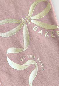Tissu rose avec un design de ruban métallique doré, avec le texte "BAKER" en relief en dessous et "BY TED BAKER" en format circulaire.
