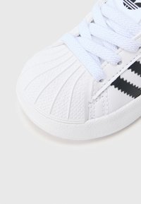 Άσπρο αθλητικό παπούτσι με υφή στη λαστιχένια μύτη, λείες δερμάτινες πλευρές και μαύρες ρίγες ως διακοσμητικά στοιχεία. Τα κορδόνια είναι γαλάζια, προσφέροντας αντίθεση.