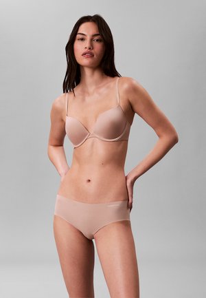 Donna in piedi con le mani sui fianchi che indossa un reggiseno color carne e biancheria intima abbinata su uno sfondo grigio semplice.