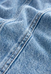  Lichtblauwe denimstof met zichtbare textuur, met een diagonale keperweef en contrasterende oranje stiksels langs de naden.