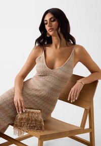 Femme aux cheveux noirs portant de grandes boucles d'oreilles dorées et une robe en tricot beige sans manches, assise sur une chaise en bois, tenant une pochette dorée à franges.