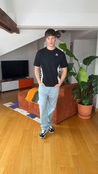 Młody mężczyzna w czarnej koszulce i jasnych dżinsach stoi w salonie z pomarańczową sofą, dużą zieloną rośliną, drewnianą podłogą i szafką pod telewizor.