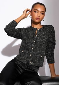 Svart, glittrande kofta med knappstängning fram och rund halsringning. Har juvel-liknande knappar och långa, texturerade ärmar. Slander svarta byxor.