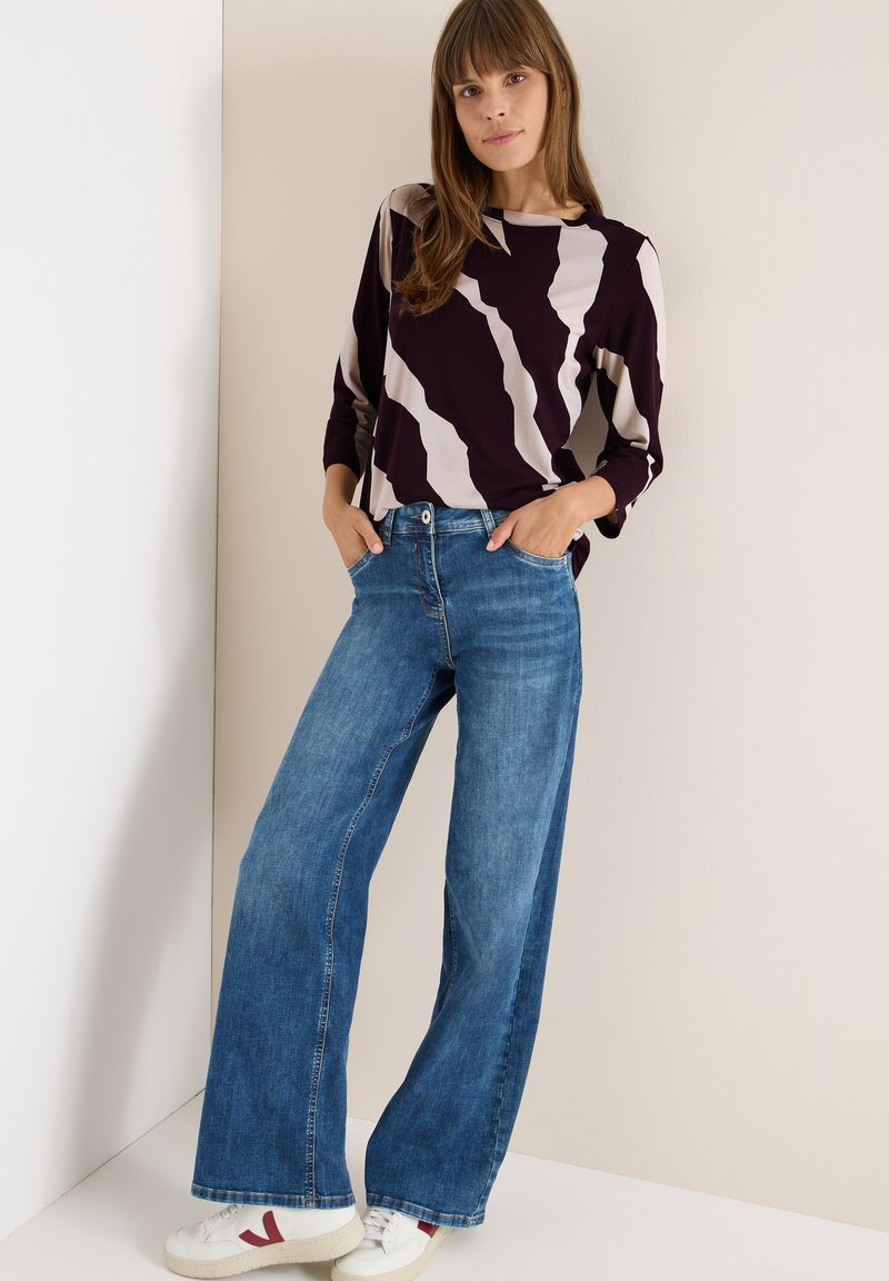 Maglietta a maniche lunghe in viola scuro con un motivo astratto in crema, abbinata a jeans a gamba larga blu. Sneakers bianche con accenti rossi.