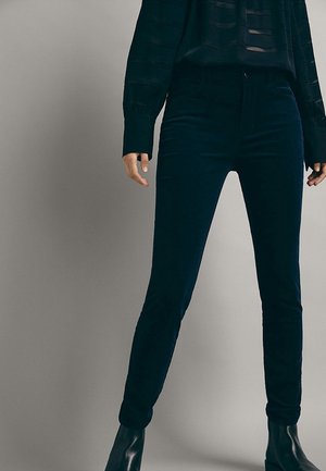 Frau trägt eng anliegende dunkle Jeans, schwarze Stiefeletten und ein langärmliges schwarzes Oberteil, steht vor neutralem Hintergrund.