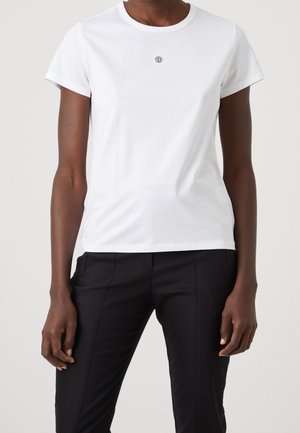 Personne portant un t-shirt blanc uni à manches courtes et un pantalon noir sur mesure, debout devant un fond neutre.