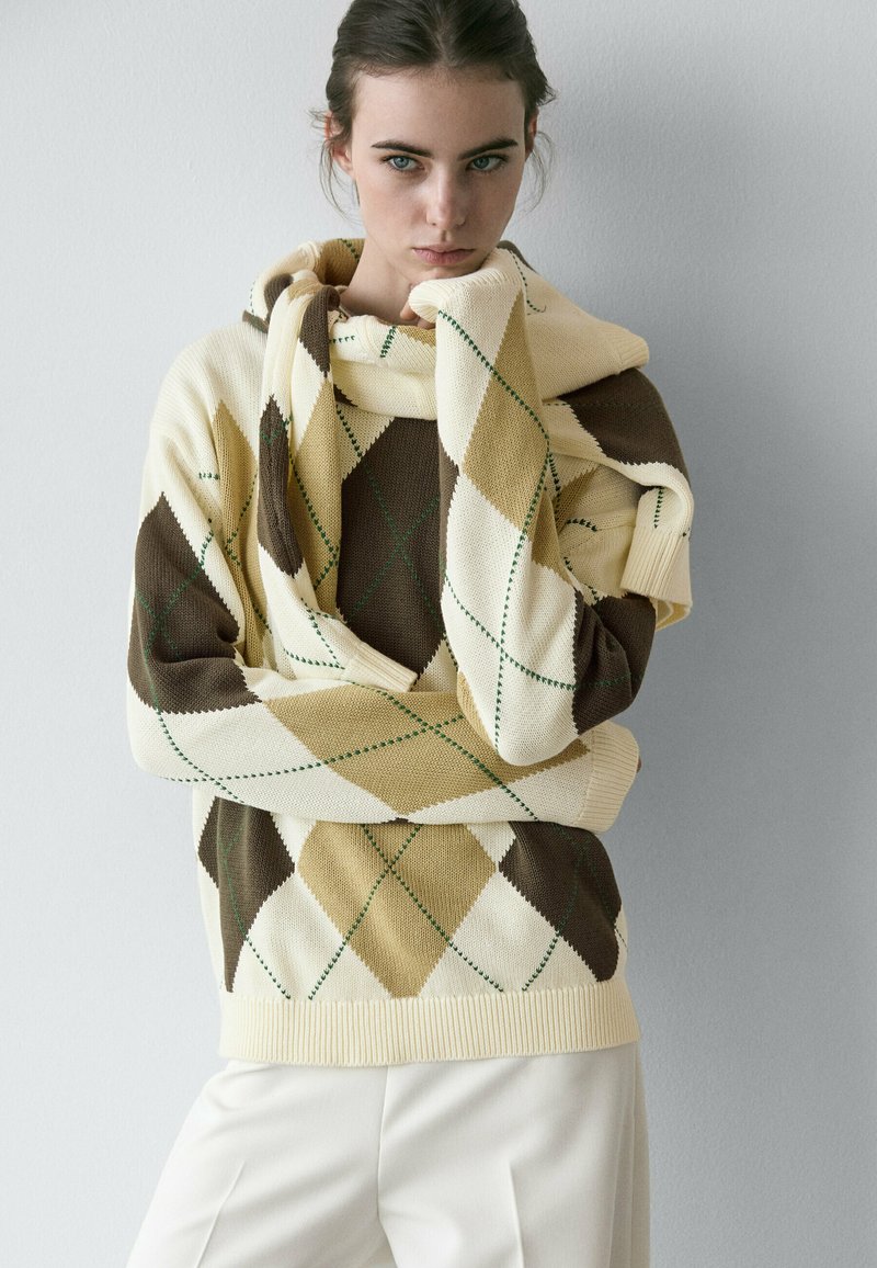 Junge Frau trägt einen cremefarbenen, braunen und beigen Argyle-Pullover mit passendem Schal, der über die Schultern gelegt ist, und steht vor einer einfarbigen hellen Wand.