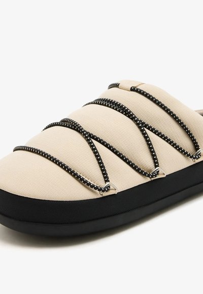 Chaussure à enfiler couleur crème avec des lacets élastiques rayés noirs et blancs qui se croisent sur la tige matelassée et une semelle noire.