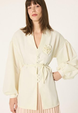 Femme portant un chemisier écru à manches longues avec des manches bouffantes, une fleur en tissu sur la poitrine, une fermeture à boutons et une ceinture à nouer à la taille.