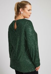 Vihreä paljetti pitkähihaisessa paidassa, jossa on avonainen selkä, rento malli ja teksturoitu pinta, varustettuna nappihaalareilla.