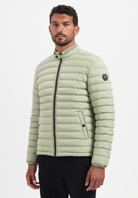 Lichtgroene pufferjas met horizontaal doorgestikt patroon, ritssluiting, hoge kraag en zeshoekige logopatch op de linkermouw.