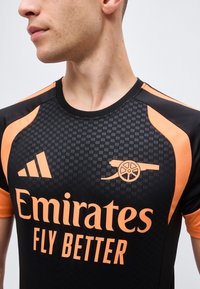 Jeune homme portant un maillot de football noir et orange avec le logo Adidas, un emblème de canon et le texte "Emirates Fly Better".
