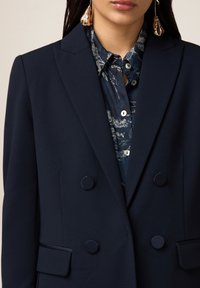 Femme portant un blazer bleu marine croisé sur une chemise foncée à motifs avec de grandes boucles d'oreilles pendantes dorées, lèvres visibles mais yeux coupés.