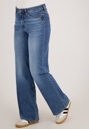 Persoon in blauwe bootcut jeans en witte sneakers met zwarte strepen, staande tegen een effen lichte achtergrond.