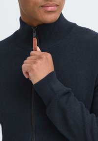 Sudadera con cremallera azul marino de cuello alto, con puños acanalados y un tirador de cremallera de cuero en contraste. Presenta una textura suave y de punto.