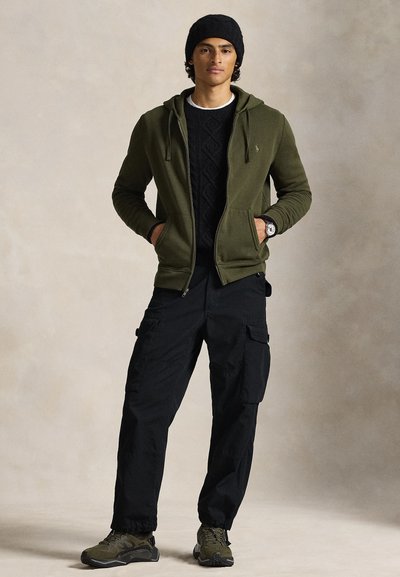 Λαδί πράσινο φούτερ με φερμουάρ, κάτω από αυτό μαύρο πλεκτό πουλόβερ με υφή, μαύρο παντελόνι cargo και σκούρα αθλητικά παπούτσια. Το μοντέλο φοράει μαύρο σκουφάκι και ρολόι καρπού.
