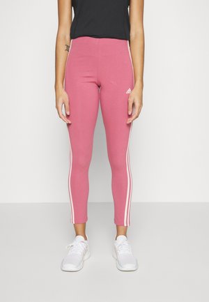 Personne portant des leggings adidas roses avec des rayures blanches sur le côté et des baskets blanches, se tenant devant un fond blanc uni.