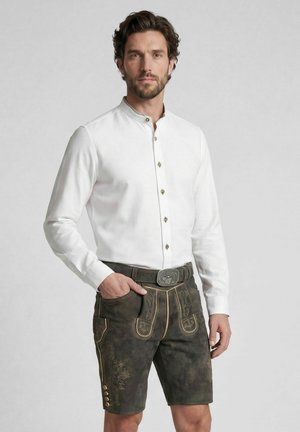 Mann trägt ein weißes langärmliges Hemd und dunkle traditionelle bestickte Lederhosen mit Gürtel, steht mit einer Hand in der Tasche vor einfachem Hintergrund.