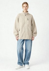 Donna che indossa una giacca beige in pile con zip e tasche, jeans larghi azzurri e sneakers bianche, in piedi davanti a uno sfondo chiaro uniforme.