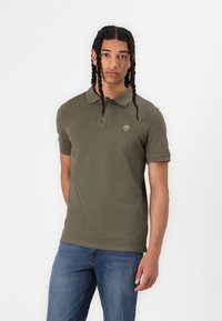 Olivgrön polo med krage och korta ärmar, prydd med en liten broderad logga på vänster bröst. Bärs med blå denimjeans.