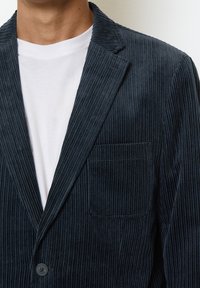 Blazer in velluto a coste blu navy con texture a coste verticali, una tasca, rever a notchs e chiusura con un solo bottone, indossato sopra una t-shirt bianca.