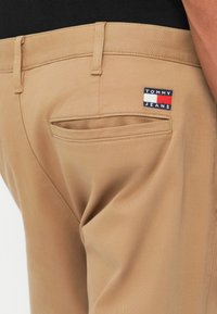 Béžové nohavice s kapsou na zadnej strane a malou nášivkou Tommy Jeans nad kapsou, nosené s čiernou košeľou.