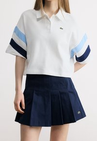 Femme portant un polo blanc court à rayures bleues et une jupe plissée bleu marine, tous deux arborant un petit logo crocodile vert.