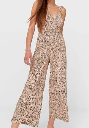 Kobieta ubrana w beżowy kombinezon z szerokimi nogawkami, wzór z czarnych nieregularnych kropek i cienkie ramiączka, stojąca na tle jednolitego jasnego tła.