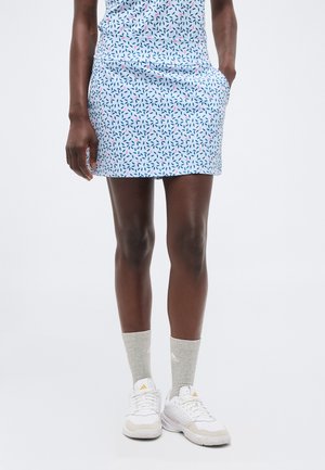 Mujer con una falda azul claro estampada, calcetines Adidas gris claro y zapatillas blancas Adidas, de pie con una mano en el bolsillo.