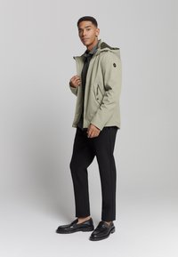 Lichtgroene waterdichte jas met een capuchon, voorzien van ritszakken en zwarte accenten, gecombineerd met zwarte broek en schoenen.