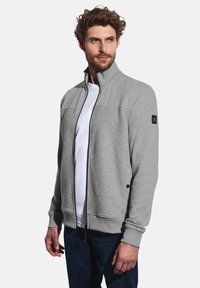 Veste zippée gris clair avec texture matelassée sur les épaules, poignets côtelés et un logo sur la manche gauche. Associée à une chemise blanche.