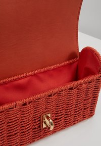 Tkaná oranžová taška ve tvaru košíku s hladkým červeným vnitřním podšívkou. Má zlatý háček na přední straně. Obdélníkový tvar.