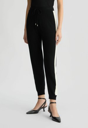 Black knit joggers featuring a drawstring waist, fitted cuffs, and contrasting white side stripes. Paired with black heeled shoes.