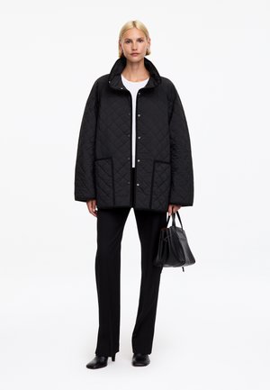 Femme portant une veste matelassée noire sur un haut blanc et un pantalon noir, tenant un sac à main noir, debout devant un fond blanc.