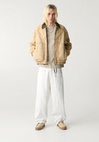 Veste tan avec des coutures visibles, manches courtes et poches avant sur une chemise beige. Pantalons larges blancs et chaussures à enfiler tan.