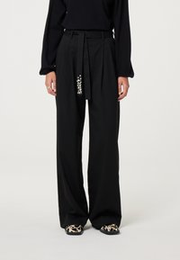 Zwarte wijde broek met een ceintuur in de taille, met een decoratief, gewatteerd detail aan één kant en een gladde stofafwerking.