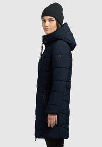 Abrigo de plumas azul marino con diseño acolchado, capucha y cremalleras laterales. El material parece suave con un acabado mate. Combinado con un gorro de punto oscuro.