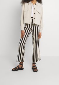 Beige cropped jasje met twee borstzakken, zwart-wit gestreepte flared broek en zwarte sandalen met gespdetails.