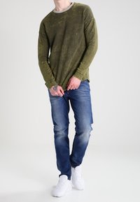 Olivgrön långärmad t-shirt med en urblekt textur, i kombination med ljusblå denimjeans. Skorna är vita sneakers.