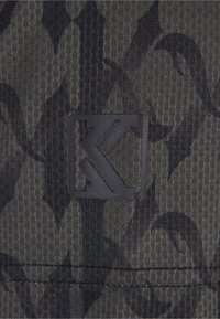 Juoda raštuota medžiaga su šešiakampiu tekstūru, dešinėje apatinėje kampoje esantis matinės juodos spalvos logotipas.