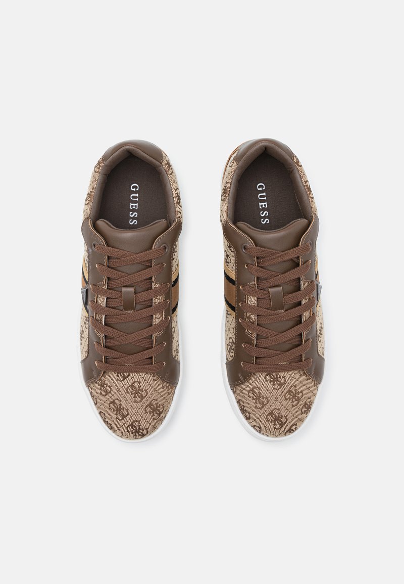 Zapatillas marrones con un patrón de logo, detalles de cuero, cordones marrones y una suela blanca. El interior es oscuro con el etiquetado de la marca.