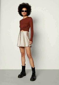 Rjava majica z dolgimi rokavi s zvinjeno sprednjo stranjo, kombinirana s sijočo, plisirano bež mini krilom. Obleko dopolnjujejo črni čevlji z vezalkami za boj.