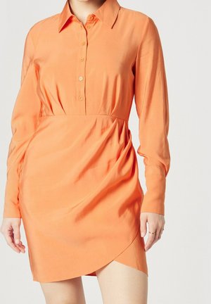 Frau trägt ein langärmeliges, orangefarbenes Kleid mit Knöpfen, einem Kragen und asymmetrischem Saum vor einem einfarbigen Hintergrund.