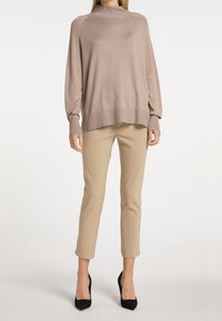 Beige gebreide trui met een hoge hals en lange mouwen, gecombineerd met beige aansluitende broek en zwarte hoge hakken.