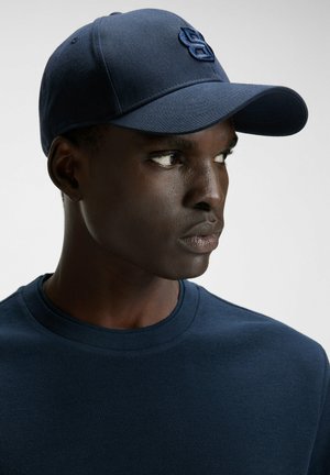 Junger Mann, der nach rechts schaut, trägt ein marineblaues, strukturiertes Hemd und eine passende Baseballkappe mit gesticktem Logo.