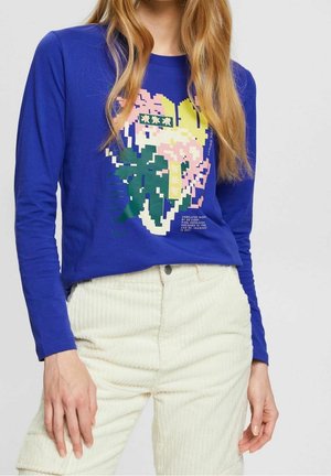 Kobieta ubrana w niebieską koszulę z długim rękawem z kolorowym, pikselowym kwiatowym wzorem oraz kremowe spodnie sztruksowe.