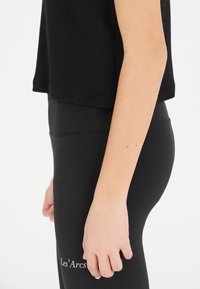 Schwarzes geripptes Cropped-Top und hochtaillierte schwarze Leggings mit weißem Schriftzug "Les'Arcs" an der Unterschenkel, glatte Textur und enge Passform.