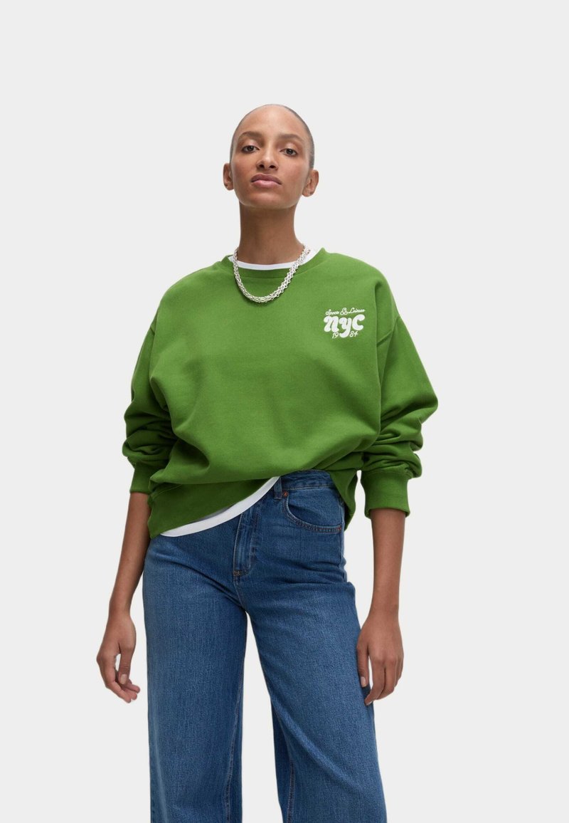 Felpa verde con collo rotondo e stampa grafica bianca "nyc" sul petto. Indossata con jeans in denim blu a vita alta. Tessuto morbido e casual.