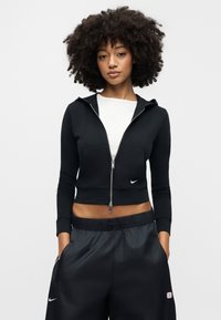 Fekete, rövidített, cipzáras kapucnis pulóver, testhezálló fazonnal, elülső zsebbel és a Nike logójával a mellkason, laza fekete sportnadrággal párosítva.